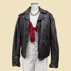 May include: A black leather motorcycle jacket with silver-toned metal zippers, a notched collar, and multiple pockets. The jacket is worn over a white top with a red and purple floral patterned scarf tied at the neck and a gold-toned chain necklace.