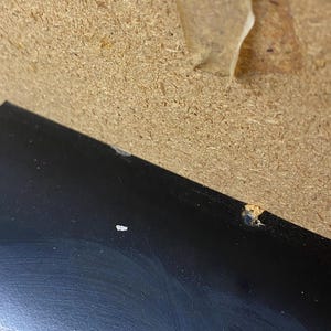 May include: A close-up shot of a black, glossy surface with minor imperfections. The surface is angled, with a light-colored, textured material in the background. There are small, light-colored specks on the black surface.