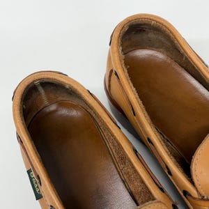 May include: A pair of tan leather boat shoes featuring a smooth brown interior lining and a small green tag with the text Bass on the side. The shoes have black lacing details around the collar and a visible stitched construction along the edges.