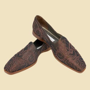 May include: A pair of brown loafers with detailed embroidery. The shoes have a square toe and a low heel. The embroidery is a lighter brown, contrasting with the darker base material. The shoes are presented against a light beige backdrop.
