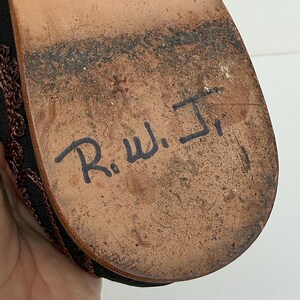 May include: Close-up of a shoe sole, showing signs of wear. The brown sole has black markings and the handwritten initials "R.W. Jr." in black ink. The shoe's upper part is visible, with a dark brown woven design.
