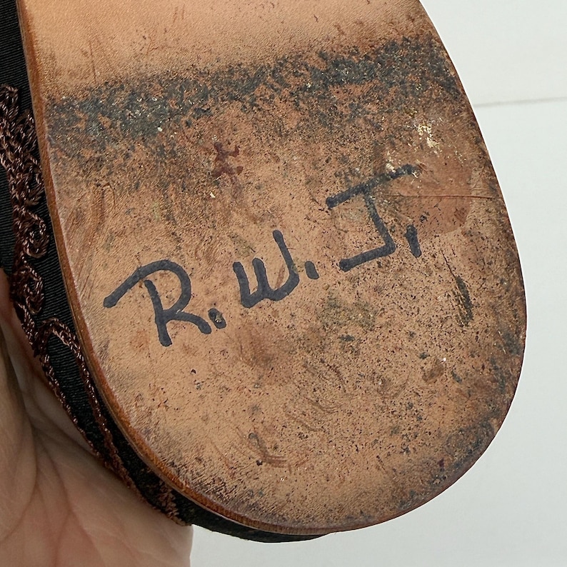 May include: Close-up of a shoe sole, showing signs of wear. The brown sole has black markings and the handwritten initials "R.W. Jr." in black ink. The shoe's upper part is visible, with a dark brown woven design.