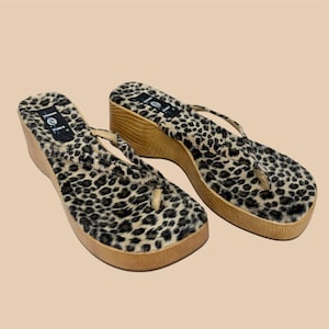 May include: A pair of platform sandals with a leopard print design. The sandals have a wooden wedge heel and a thong strap. The brand name "lei" is visible on the top of the sandals.