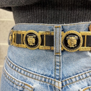 Puede incluir: Un cintur&oacute;n de cuero negro con herrajes de metal dorado que presentan un patr&oacute;n de greca repetitivo y medallones circulares. Cada medall&oacute;n muestra un rostro dorado en relieve con un dise&ntilde;o de casco alado sobre un fondo de esmalte negro.