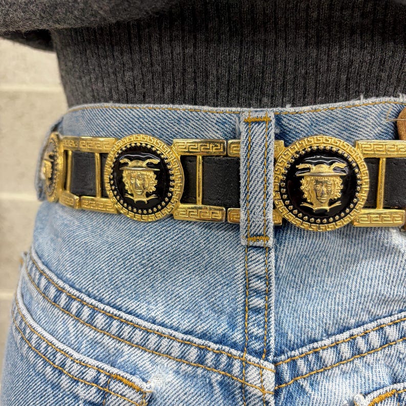 Puede incluir: Un cintur&oacute;n de cuero negro con herrajes de metal dorado que presentan un patr&oacute;n de greca repetitivo y medallones circulares. Cada medall&oacute;n muestra un rostro dorado en relieve con un dise&ntilde;o de casco alado sobre un fondo de esmalte negro.