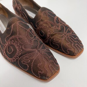 May include: A pair of brown slip-on shoes with square toes. The shoes feature an intricate embroidered design in a darker brown thread. The shoes have a low heel and a smooth, polished finish, suitable for a formal or semi-formal event.