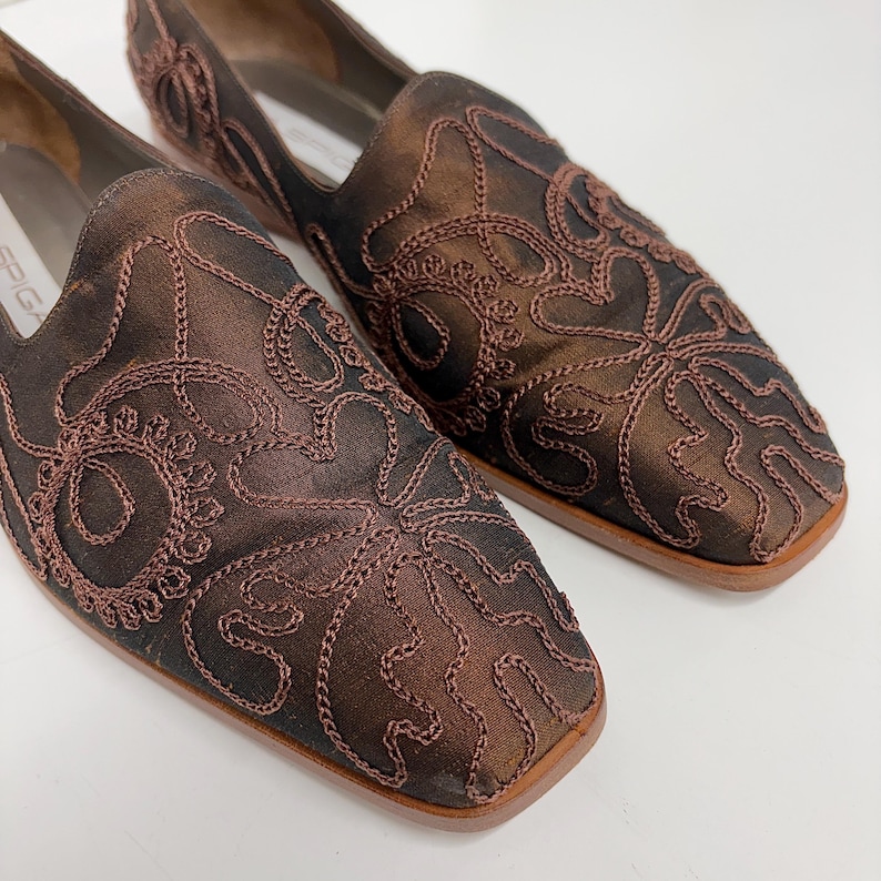 May include: A pair of brown slip-on shoes with square toes. The shoes feature an intricate embroidered design in a darker brown thread. The shoes have a low heel and a smooth, polished finish, suitable for a formal or semi-formal event.