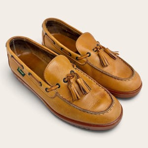 May include: A pair of tan leather loafers featuring a moccasin-style toe, decorative tassels on the vamp, and a lacing detail around the collar. The shoes have a small green tag with the word Bass on the side and a dark brown sole.