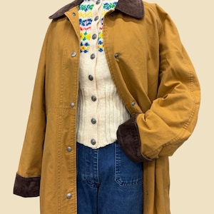 May include: A mustard yellow long coat with a dark brown corduroy collar and cuffs, featuring silver snap buttons. Underneath is a cream-colored knit cardigan with a colorful floral pattern around the neckline and blue denim jeans.