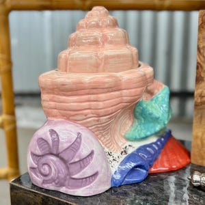 May include: A ceramic decorative object shaped like a cluster of seashells. The central shell is light pink, featuring a tiered, spiral structure. It is accented by a purple shell at the base, a bright turquoise shell on the side, and a red base.