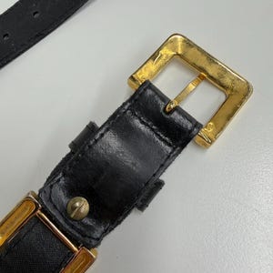 Puede incluir: Un cintur&oacute;n negro con una hebilla cuadrada de metal dorado y una trabilla de metal dorado a juego. La correa del cintur&oacute;n est&aacute; hecha de un material negro con textura visible y est&aacute; asegurada con un peque&ntilde;o remache de metal dorado.