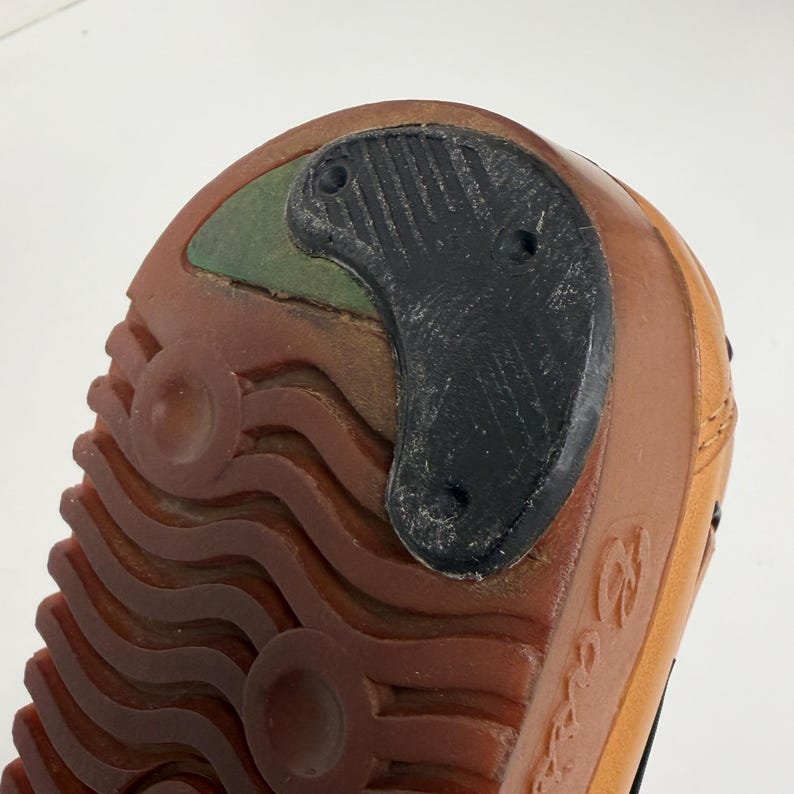 May include: The underside of a shoe featuring a brown rubber sole with a wavy tread pattern and circular indentations. A black, crescent-shaped rubber heel tap is attached to the back, partially covering a small green section of the sole.