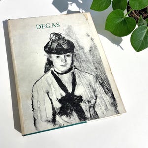 May include: A hardcover book titled DEGAS with a white cover featuring a black and white charcoal-style illustration of a person wearing a hat and a dark necktie. The book has a rectangular shape with visible wear on the top right corner edge.