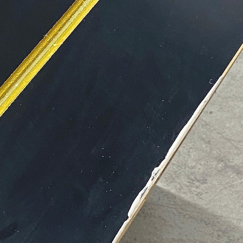 May include: Close-up of a black surface with a gold and white stripe. The edge of the object is visible, showing a white border with some damage. The surface appears to be smooth and reflective.