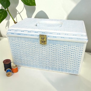 May include: A vintage sewing box with a light blue and white woven pattern. The box has a handle on the top and a gold-colored clasp. Several spools of thread in various colors are next to the box.