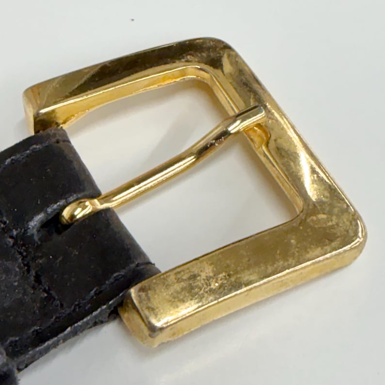 Puede incluir: Un cintur&oacute;n marr&oacute;n oscuro con una hebilla rectangular de metal dorado con acabado pulido. La correa del cintur&oacute;n est&aacute; hecha de un material texturizado y se asegura mediante un perno que pasa a trav&eacute;s del marco de la hebilla para cerrarlo.