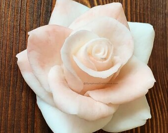 May include: A delicate, hand-crafted, white and pink rose soap. The soap is shaped like a rose with a swirl pattern in the center. The soap is on a brown wooden surface.