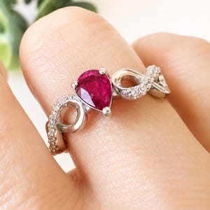 May include: A silver-toned ring featuring a central pear-shaped deep pink gemstone held by prongs. The band has an infinity-style design accented with small, clear, sparkling stones embedded along the metal surface on both sides of the center stone.