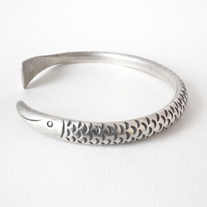 May include: A silver-toned cuff bracelet with a fish design. The bracelet features a fish head at one end and a tail-like shape at the other. The body of the fish is detailed with scale patterns. The bracelet is open and adjustable.