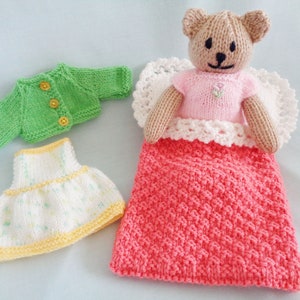 May include: A knitted teddy bear wearing a pink dress and a white crocheted halo, lying in a coral knitted blanket. The bear is surrounded by a green knitted cardigan and a white and yellow knitted skirt.