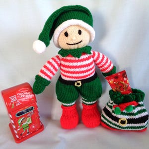 May include: A knitted elf doll wearing a green hat, red and white striped top, green trousers, and red boots. The elf is next to a red postbox and holds a small bag. A small bag with a black and white stripe pattern is also present.