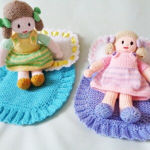 May include: Two knitted dolls with brown and blonde hair are lying on blue and purple knitted blankets. The dolls are wearing striped dresses and have embroidered faces.