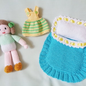 May include: A knitted doll with brown hair, a green and white shirt, and pink legs. The doll is wearing a yellow and green striped dress and is lying in a blue knitted sleeping bag with a white pillow and yellow trim.