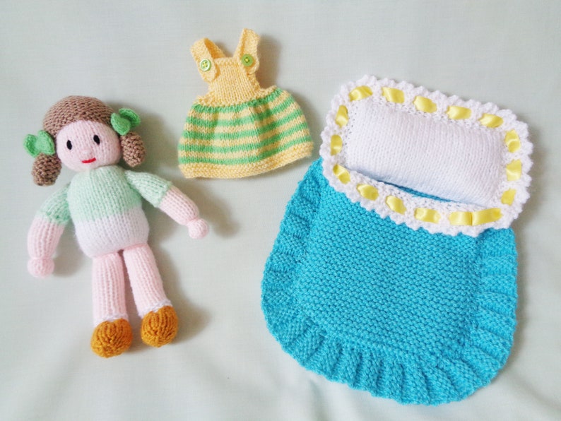 May include: A knitted doll with brown hair, a green and white shirt, and pink legs. The doll is wearing a yellow and green striped dress and is lying in a blue knitted sleeping bag with a white pillow and yellow trim.