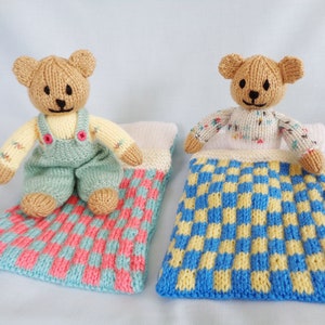 Peut inclure: Deux ours en peluche tricotés, l'un assis et l'autre couché dans un sac de couchage tricoté. Le sac de couchage a un motif à carreaux bleu et jaune. L'ours en peluche assis porte une salopette et a un sac de couchage à carreaux avec des carrés roses et bleus.