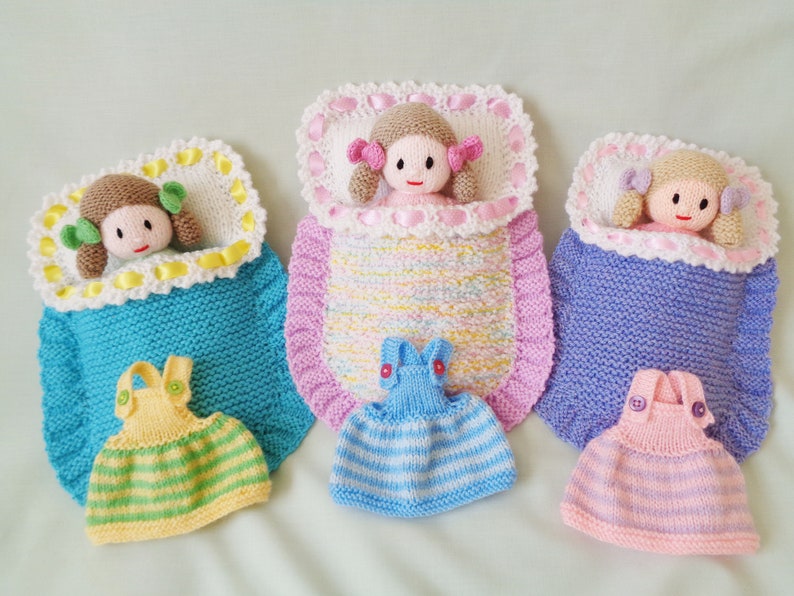 May include: Three knitted dolls in sleeping bags with matching knitted dresses. The dolls have pink cheeks and brown hair with pink bows. The sleeping bags are blue, pink, and purple with white trim. The dresses are yellow and green, blue and white, and pink and purple.