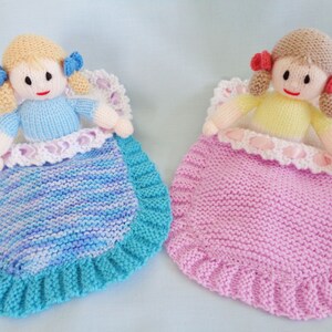 May include: Two knitted dolls in sleeping bags. The doll on the left has blonde hair, a blue dress, and a blue and white striped sleeping bag. The doll on the right has brown hair, a yellow dress, and a pink sleeping bag.