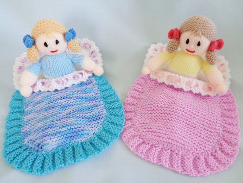 May include: Two knitted dolls in sleeping bags. The doll on the left has blonde hair, a blue dress, and a blue and white striped sleeping bag. The doll on the right has brown hair, a yellow dress, and a pink sleeping bag.