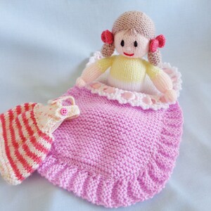May include: A knitted doll with brown hair and a pink dress is lying in a pink knitted sleeping bag. The doll is wearing a yellow and white striped dress.