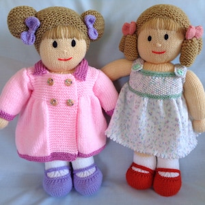 May include: Two handmade knitted dolls. One doll wears a pink coat with gold buttons, purple shoes, and a brown hair with purple bows. The other doll wears a white dress with a floral pattern, red shoes, and brown hair with pink bows.