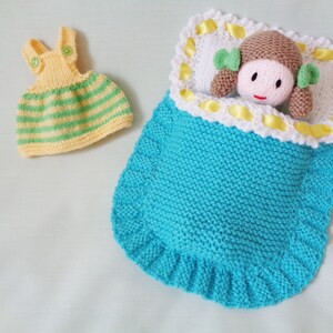 May include: A knitted doll with brown hair and a yellow and green striped dress is sleeping in a blue knitted sleeping bag with a white and yellow trim.