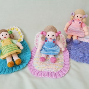 May include: Three knitted dolls with different colored dresses and matching knitted blankets. The dolls are wearing yellow, blue, and pink dresses. The blankets are blue, pink, and purple.