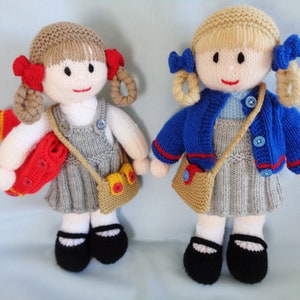 Peut inclure: Deux poupées en tricot habillées d'uniformes scolaires. Une poupée porte un cardigan bleu et l'autre une robe à tablier grise. Les deux poupées ont les cheveux bruns et tiennent des sacs.