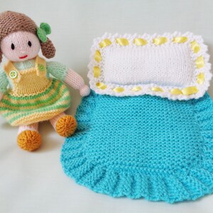 May include: A knitted doll with brown hair, a green and yellow dress, and a button on her chest, standing next to a blue knitted blanket with a white pillow and yellow trim.