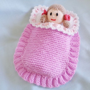 May include: A pink knitted doll sleeping bag with a white crocheted trim and a doll with brown hair and a pink and white dress inside.