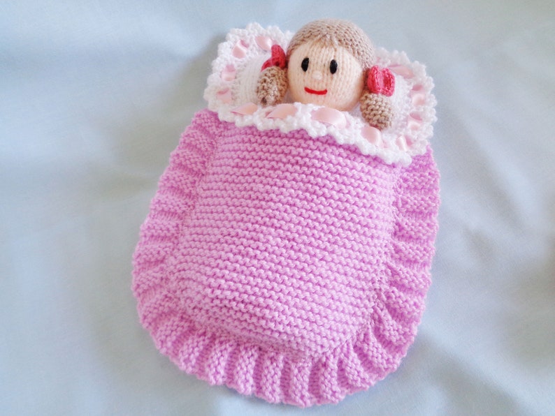 May include: A pink knitted doll sleeping bag with a white crocheted trim and a doll with brown hair and a pink and white dress inside.