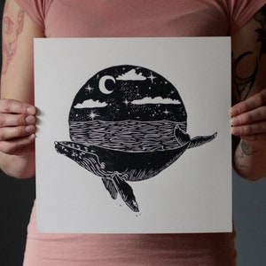 May include: Black and white linocut print of a whale swimming through a circle with a crescent moon, stars, and clouds. The whale is silhouetted against the sea and sky.