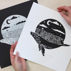 May include: Black and white whale art print. The print features a whale swimming under a night sky with a crescent moon, stars, and clouds. The art is displayed on a white background, with a black print in the background.