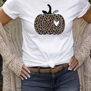 以下が含まれることがあります： 白いTシャツに、黒と茶色のレオパード柄の南瓜が描かれており、白いハートが描かれています。シャツはウエストで結ばれており、人はベージュのカーディガンを着ています。
