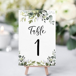 May include: White table number card with green watercolor foliage and the text "Table 1" in black calligraphy.