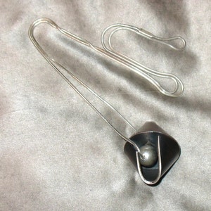 Puede incluir: Un collar de cadena de plata con un colgante negro que contiene una esfera de plata en su interior. El colgante tiene la forma de un tri&aacute;ngulo plegado.