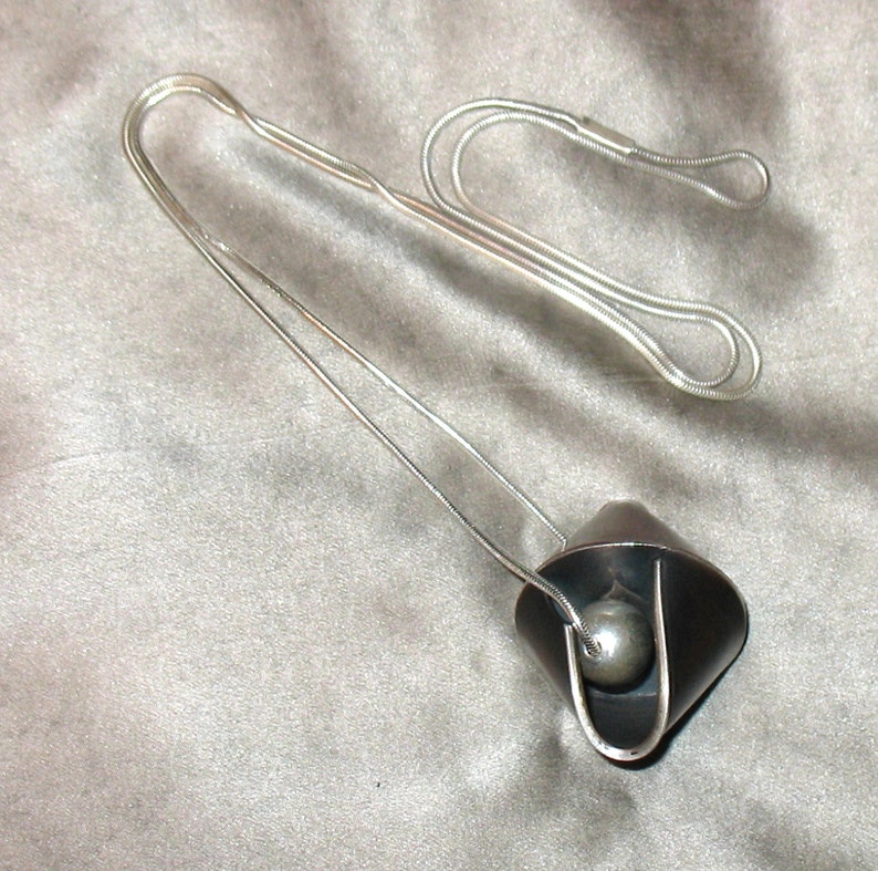 Puede incluir: Un collar de cadena de plata con un colgante negro que contiene una esfera de plata en su interior. El colgante tiene la forma de un tri&aacute;ngulo plegado.