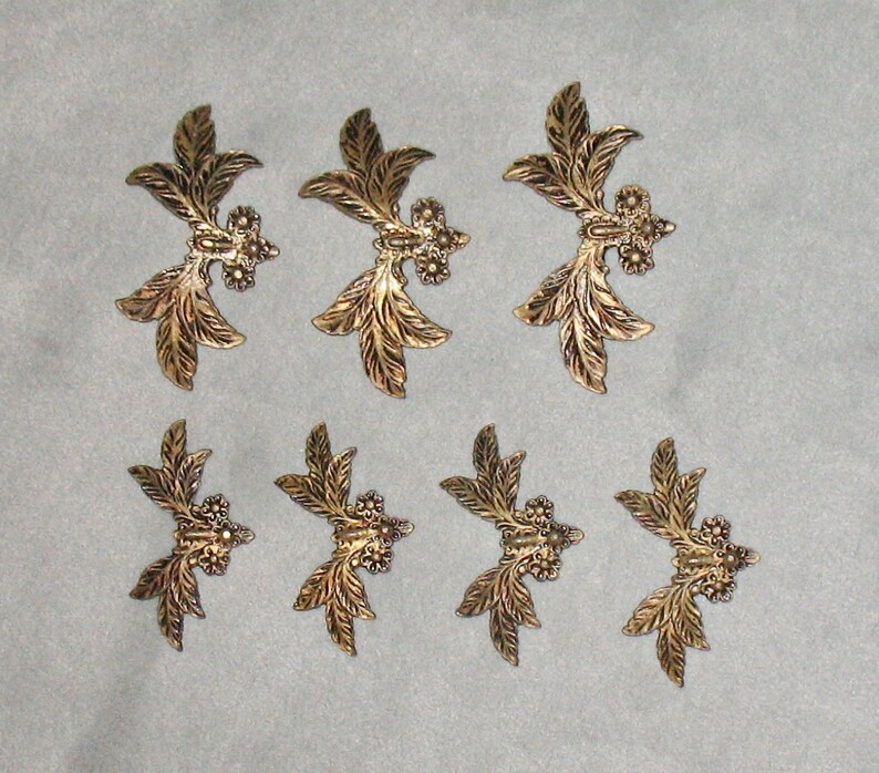 May include: Set of eight gold-toned metal leaf-shaped embellishments. Each embellishment features a central design of three small circles and a raised, textured leaf pattern.