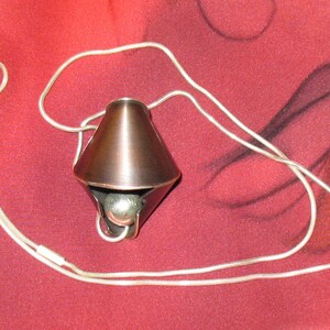 Puede incluir: Un colgante de color cobre con una bola de plata en su interior, colgando de una cadena de plata. El colgante tiene forma de cono con una punta afilada y una base ancha. La cadena est&aacute; hecha de peque&ntilde;os anillos unidos.