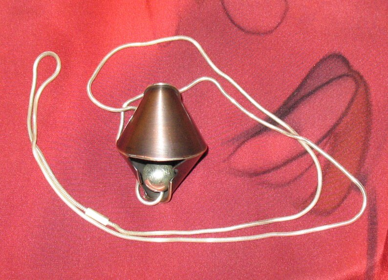 Puede incluir: Un colgante de color cobre con una bola de plata en su interior, colgando de una cadena de plata. El colgante tiene forma de cono con una punta afilada y una base ancha. La cadena est&aacute; hecha de peque&ntilde;os anillos unidos.