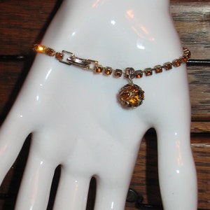 May include: A gold tone bracelet with a dangling amber colored rhinestone ball charm. The bracelet is made of small square rhinestones.